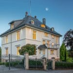 Historische Stadtvilla bei Dämmerung mit beleuchteten Fenstern und schmiedeeisernem Zaun