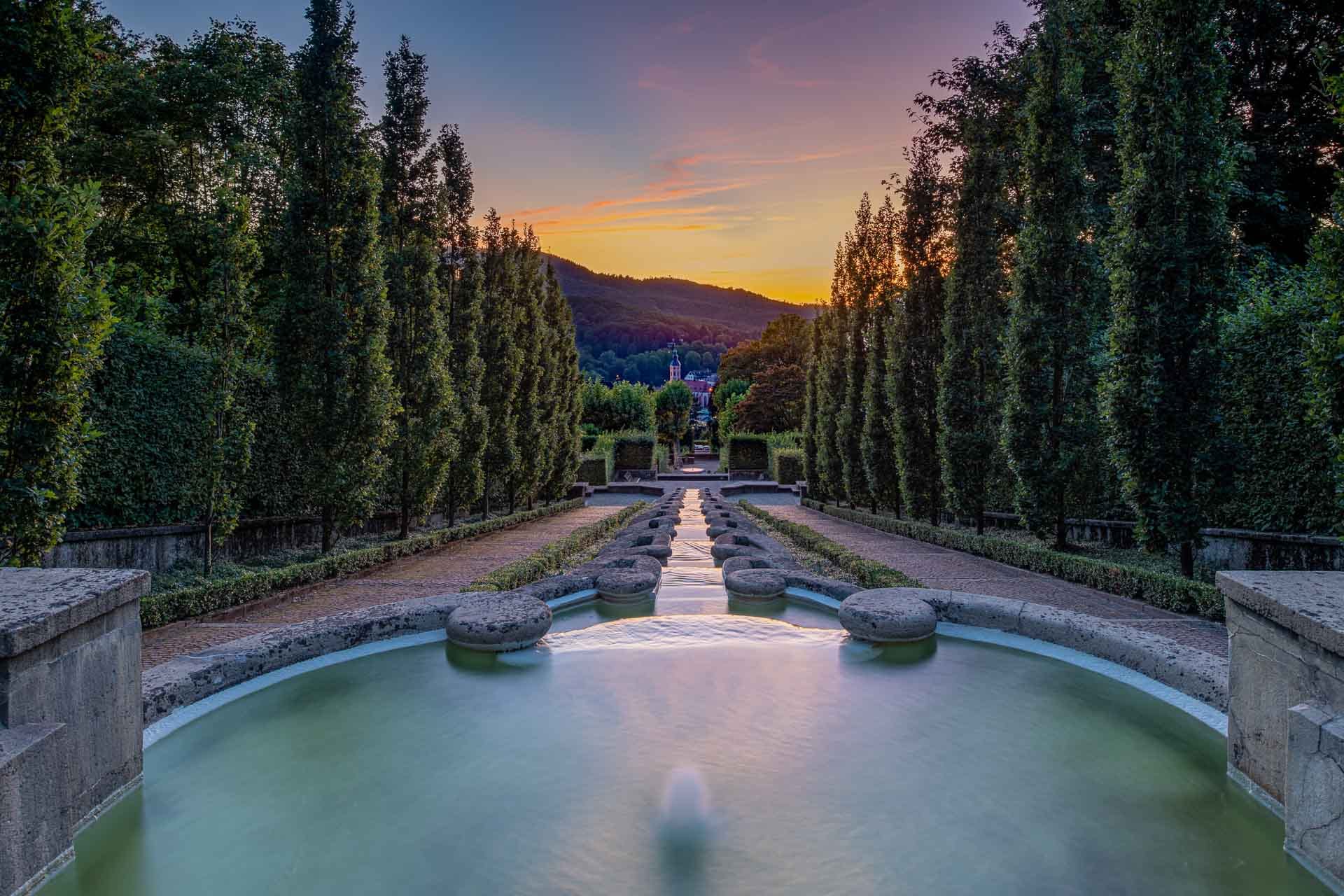 Langzeitbelichtung der Wasserspiele im Paradiesgarten Baden-Baden mit symmetrischer Baumreihe und Sonnenuntergang im Hintergrund